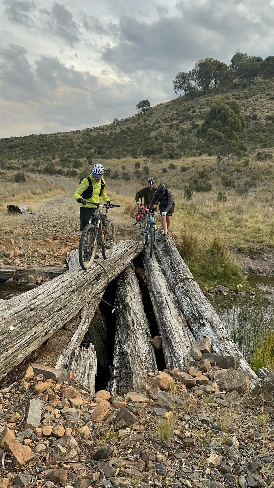 Three helmeted riders guiding their bikes on foot across a weathered log bridge.