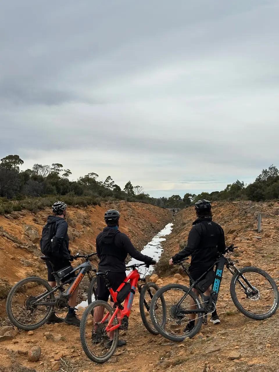 Three riders standing with their Roar bikes beside a rocky channel in Tasmania.